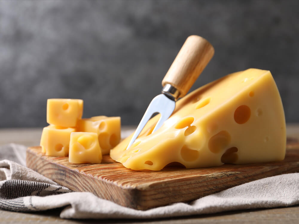 Emmentaler-Stück mit typischen Löchern und Käsegabel auf einem Holzbrett – nussiger Käse zum Überbacken von Gratins und Aufläufen