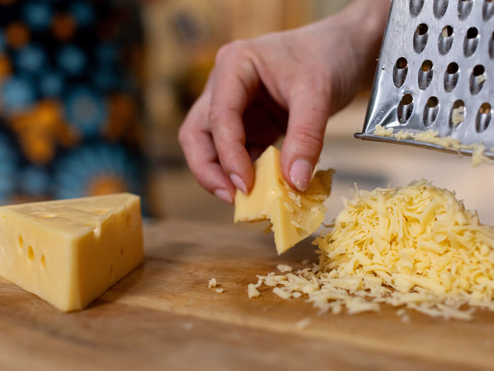 Hand reibt ein Stück Käse mit der Käsereibe auf einem Schneidebrett – frisch geriebener Käse zum Überbacken von Aufläufen