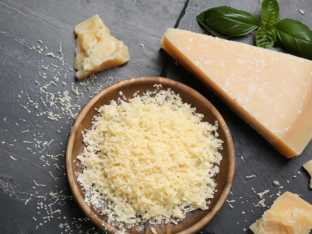Geriebener Parmesan in einer Holzschale mit Käsestücken und Basilikum – intensiver Käse als Finish zum Überbacken
