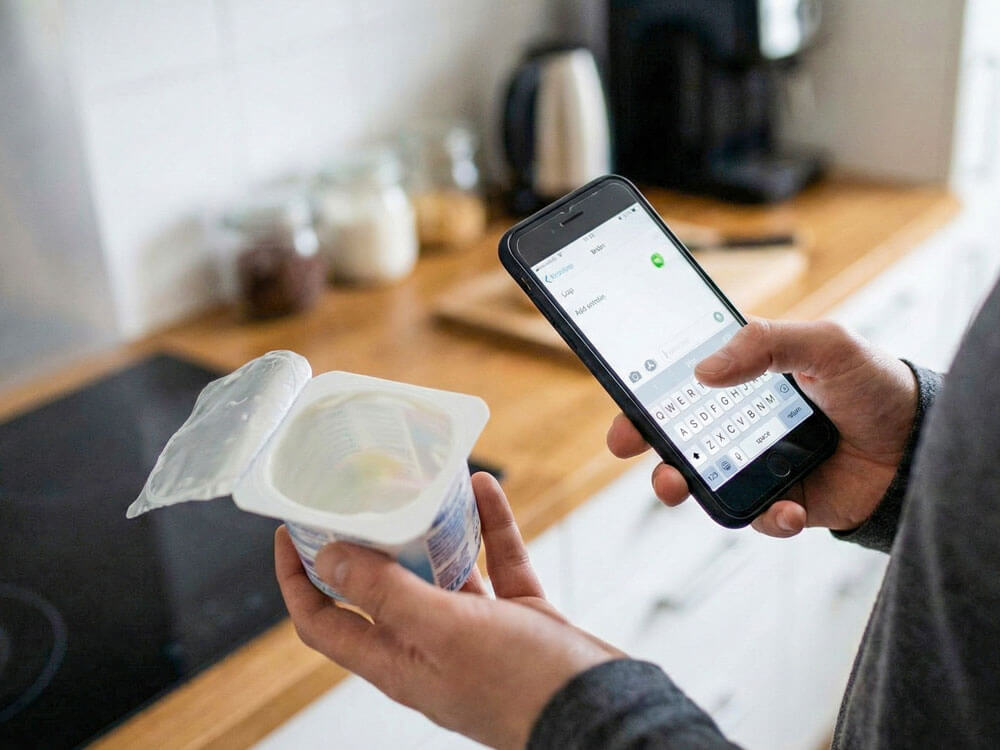 Hand tippt fehlenden Joghurt in Einkaufslisten-App ein – Spielregel für digitalen Basisvorrat und Küchen-Lifehack gegen fehlende Zutaten