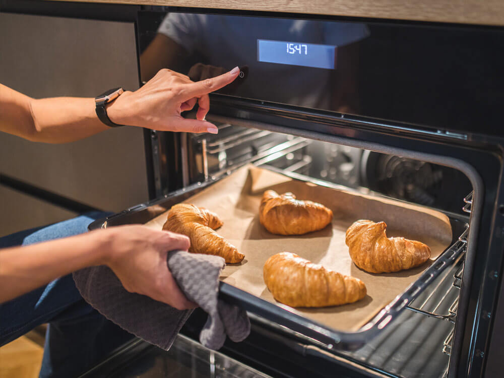 Frau nimmt Blech mit goldbraunen Croissants aus dem Backofen, gebacken mit Umluft