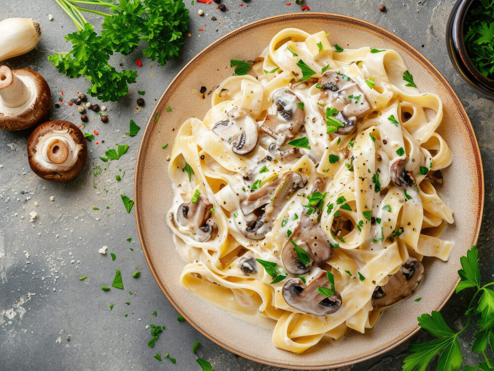 Pilz-Pasta mit cremiger Soße und Petersilie auf einem Teller, daneben Champignons und Knoblauch