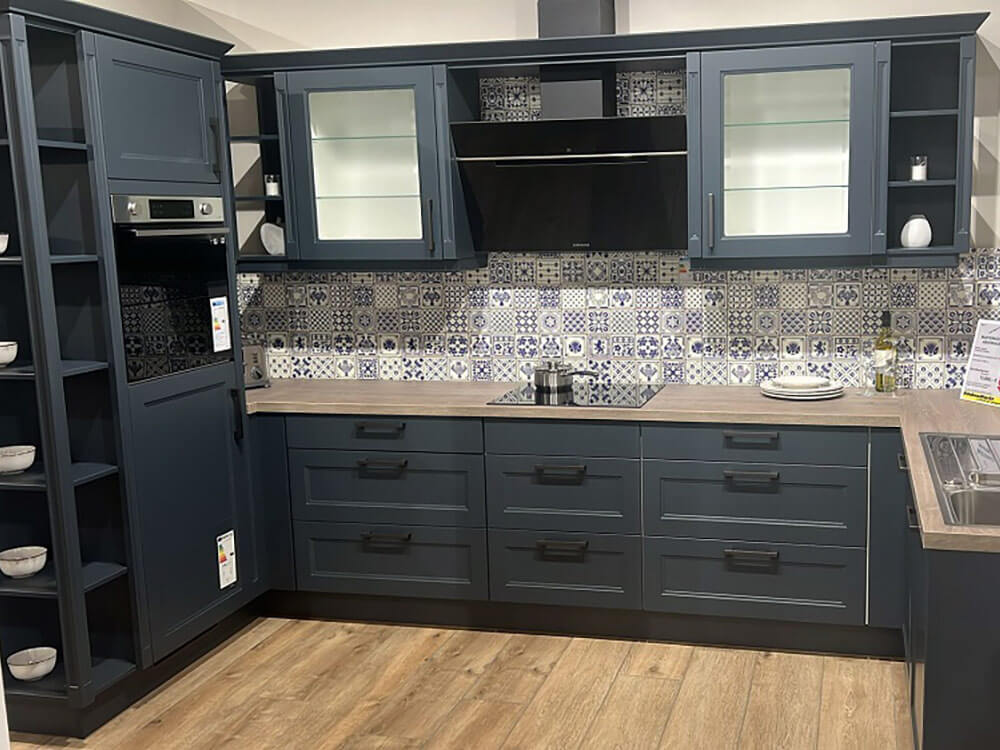 Gesamtansicht der U-Küche Landhaus Cascada (Koje 17) in Dunkelblau mit Kochfeld, Dunstabzug und gemusterten Fliesen.
