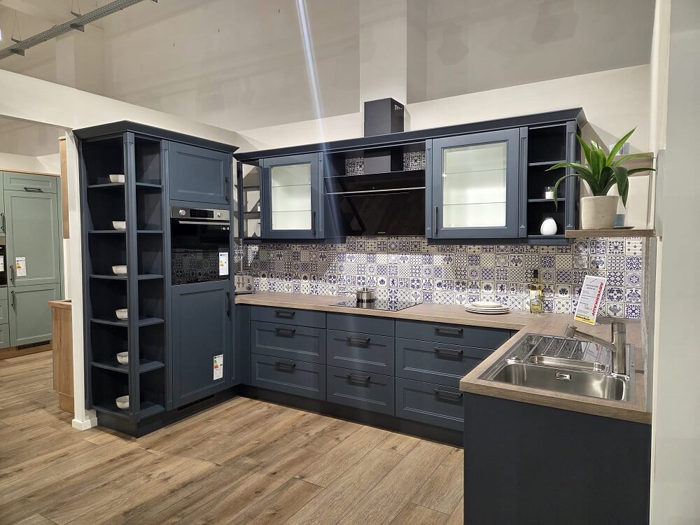 Weitwinkel-Ansicht der U-Küche Landhaus Cascada (Koje 17) mit hohem Regal, Kochbereich und Spüle in der Ausstellung.