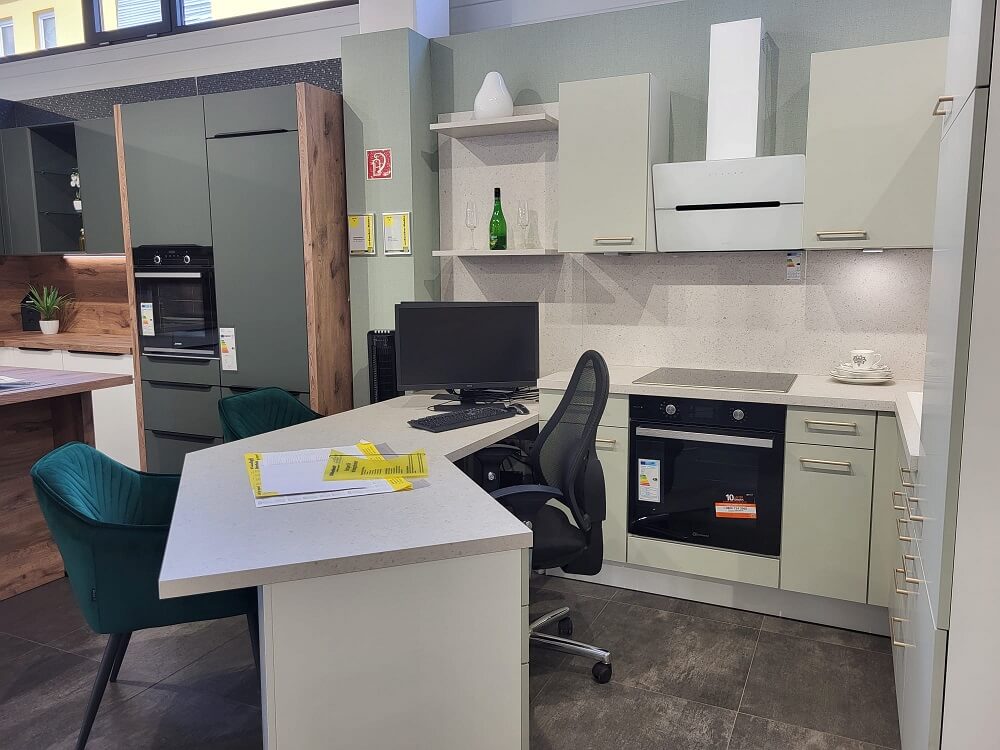 Ausstellungsküche in L‑Form mit hellgrünen Fronten, weißer Arbeitsplatte, Dunstabzugshaube und integriertem Computerarbeitsplatz mit Stuhl und Monitor