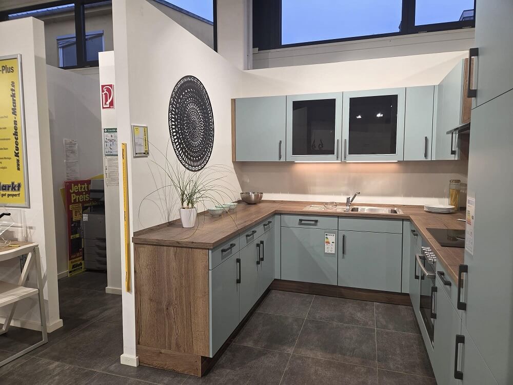 U-Küche in modernem Design mit Glasoberschränken, Holzrückwand und hellblauen Fronten – Ausstellungsküche Touch bei KüchenMarkt Landshut Nord.